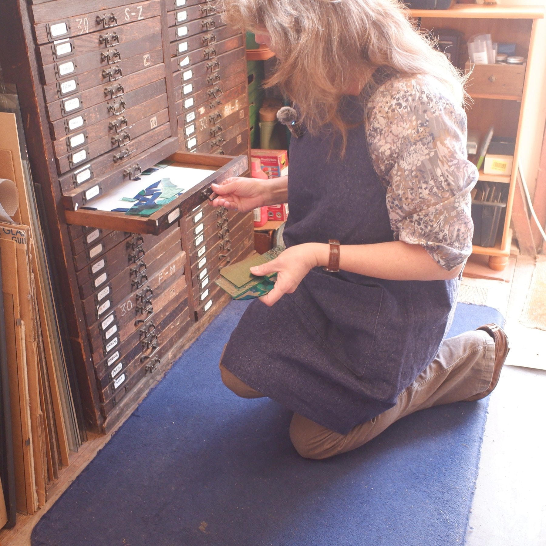 Mary Corkery kneeling at an old wooden plan chest in her workshop, taking a sheet of glass from a drawer