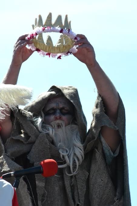 Statue of bearded wizard holding aloft a crown decorated with pink and purple flowers, against clear blue sky.