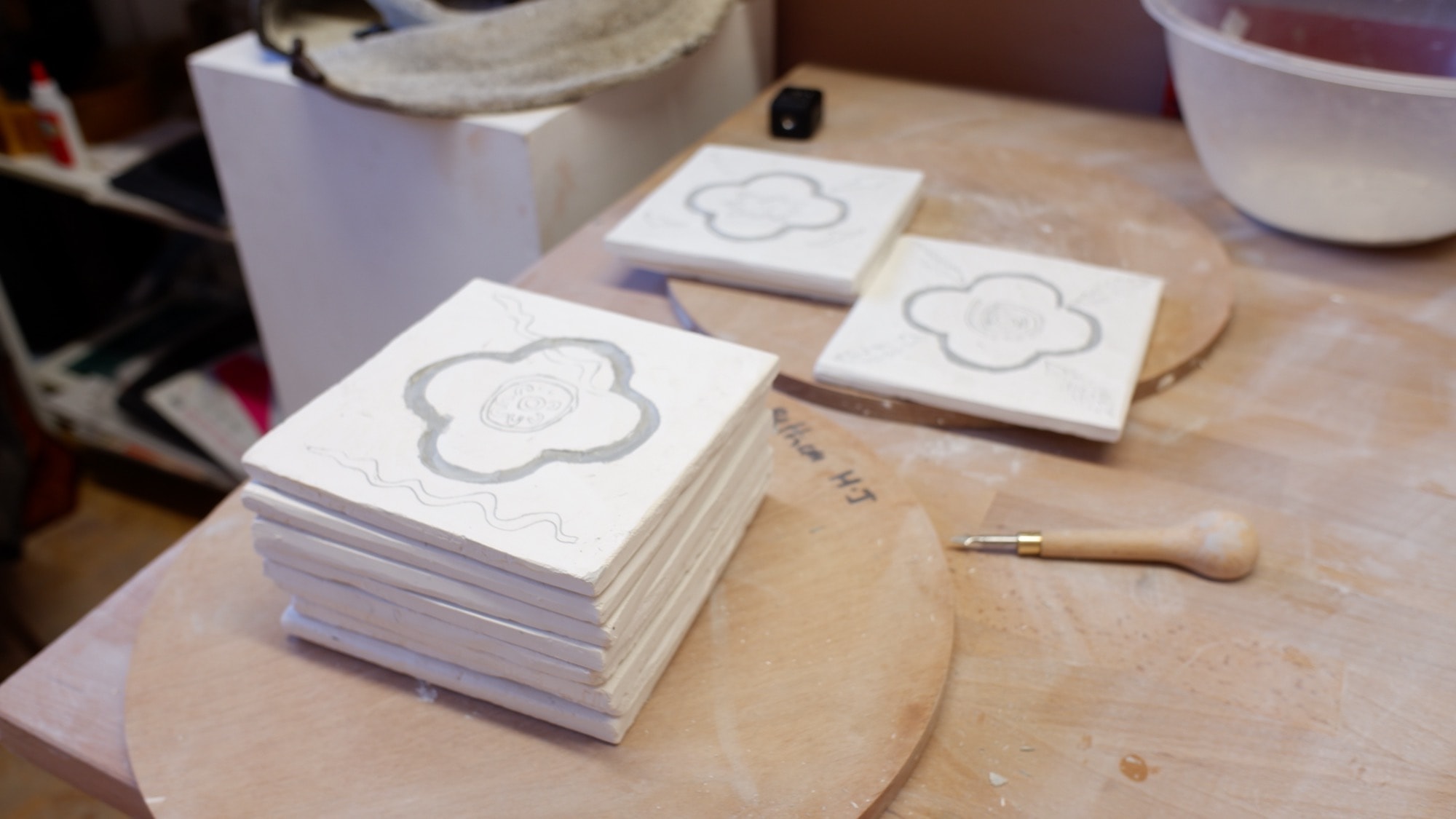 A stack of stamped porcelain tiles drying on a wooden board, with more tiles on the table behind
