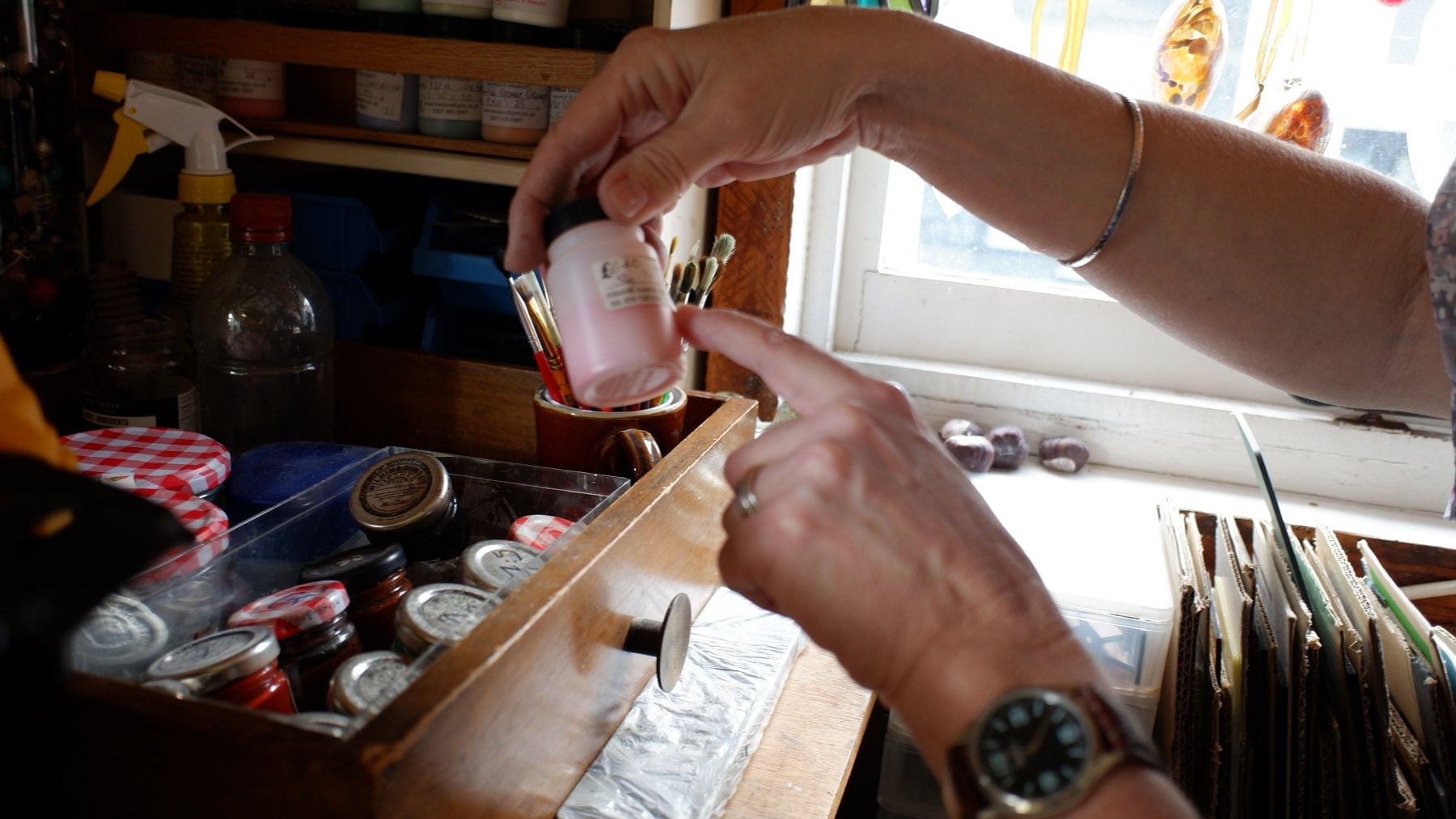 Hands taking brushes from a pot beside jars of glass paint by a window