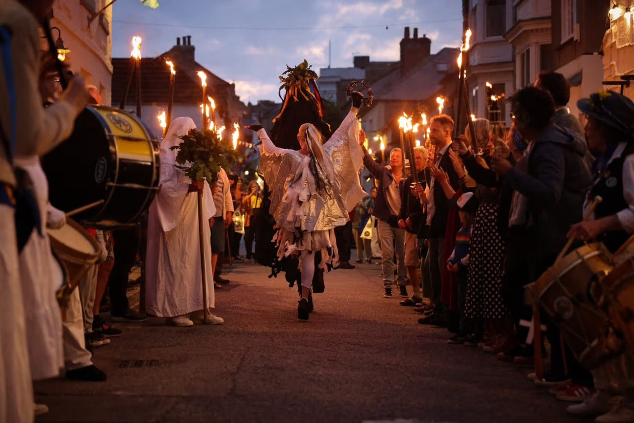 Figures in white robes and wings parading down a street lined with flaming torches at dusk, Georgian brick buildings behind,