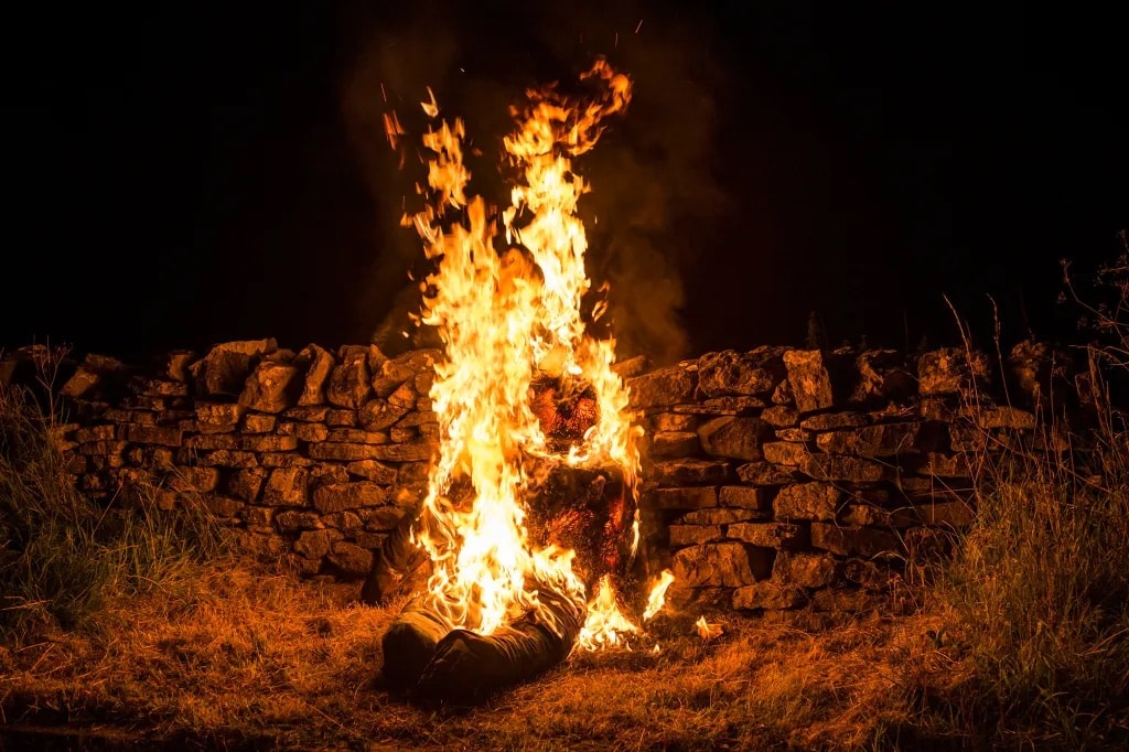 Bright bonfire with orange and yellow flames rising against a dark night sky, stone wall and dry grass visible in background.