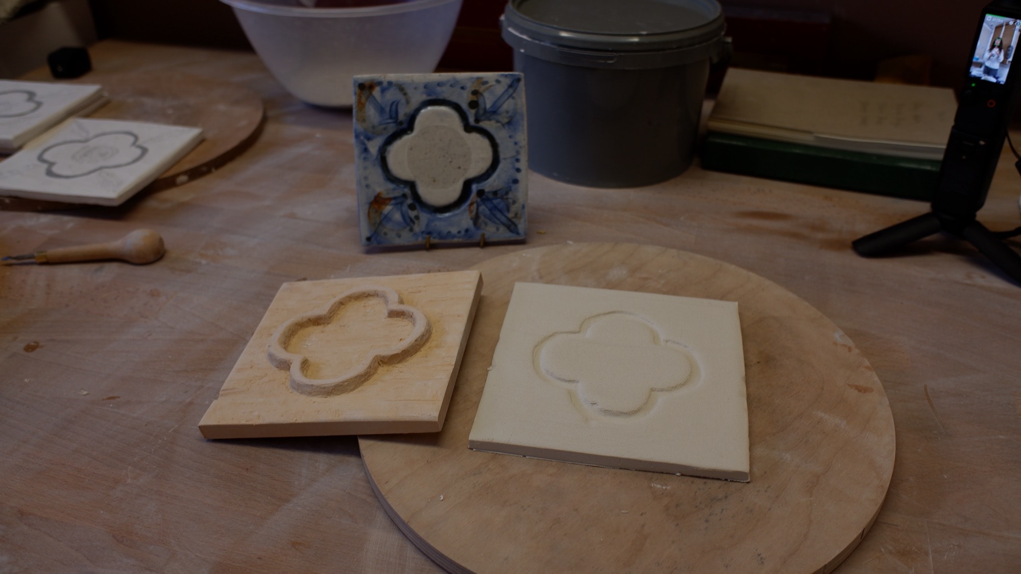 A hand-carved wooden quatrefoil stamp beside the porcelain tile it has been pressed into, with a finished blue tile behind