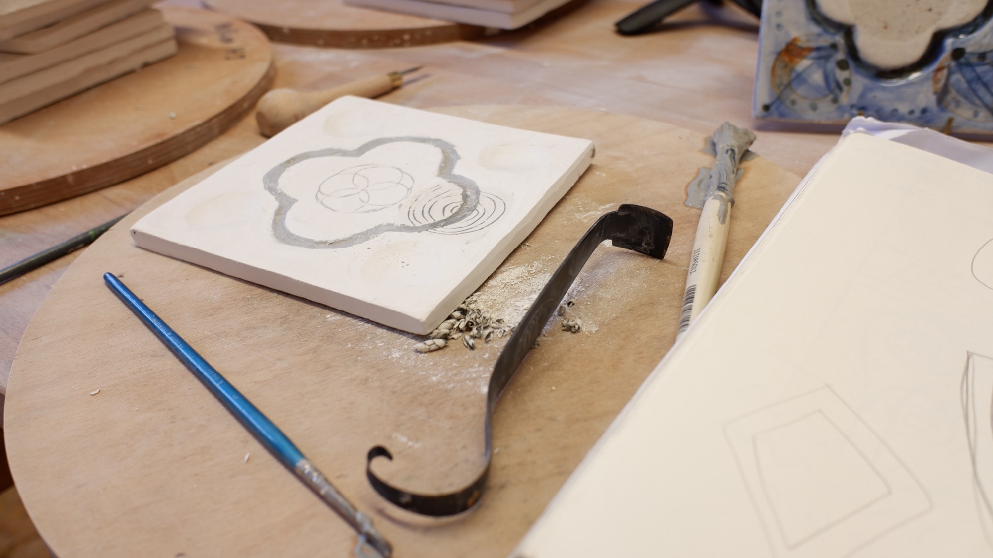 A stamped porcelain tile with a quatrefoil outline in grey slip, a carving tool and brush beside it