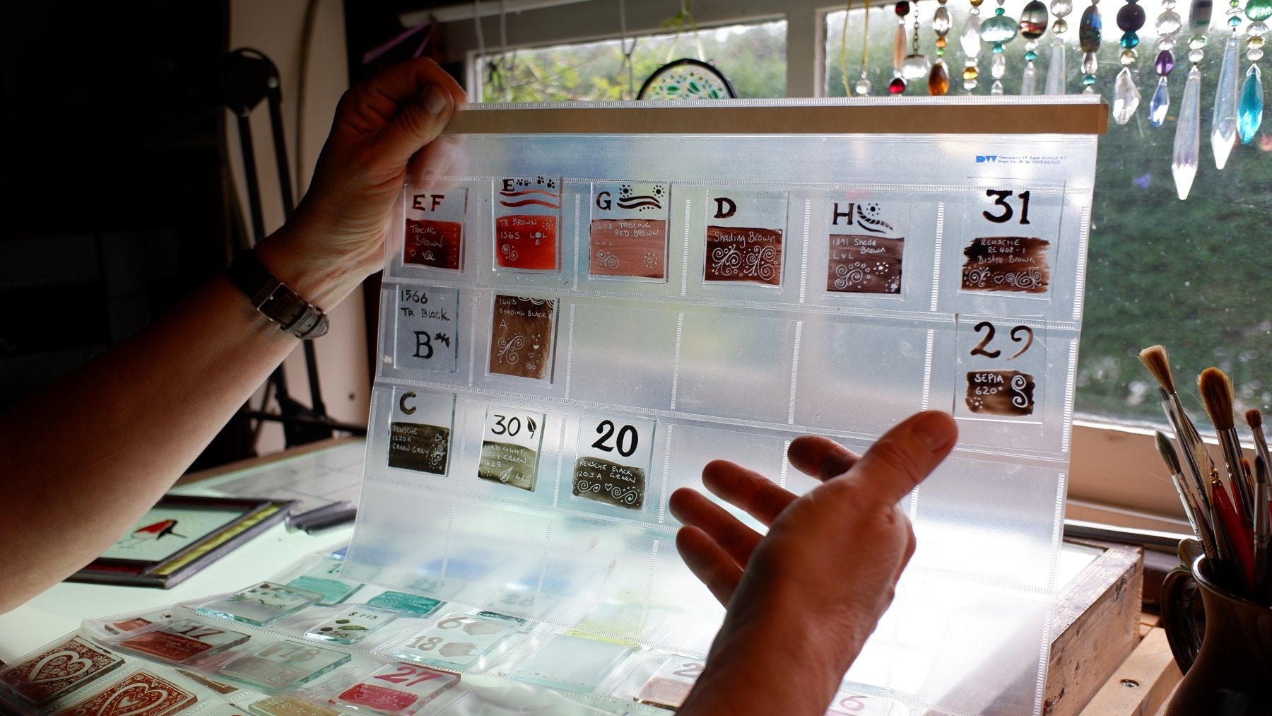 Glass paint test tiles in numbered pockets, held up against the workshop window