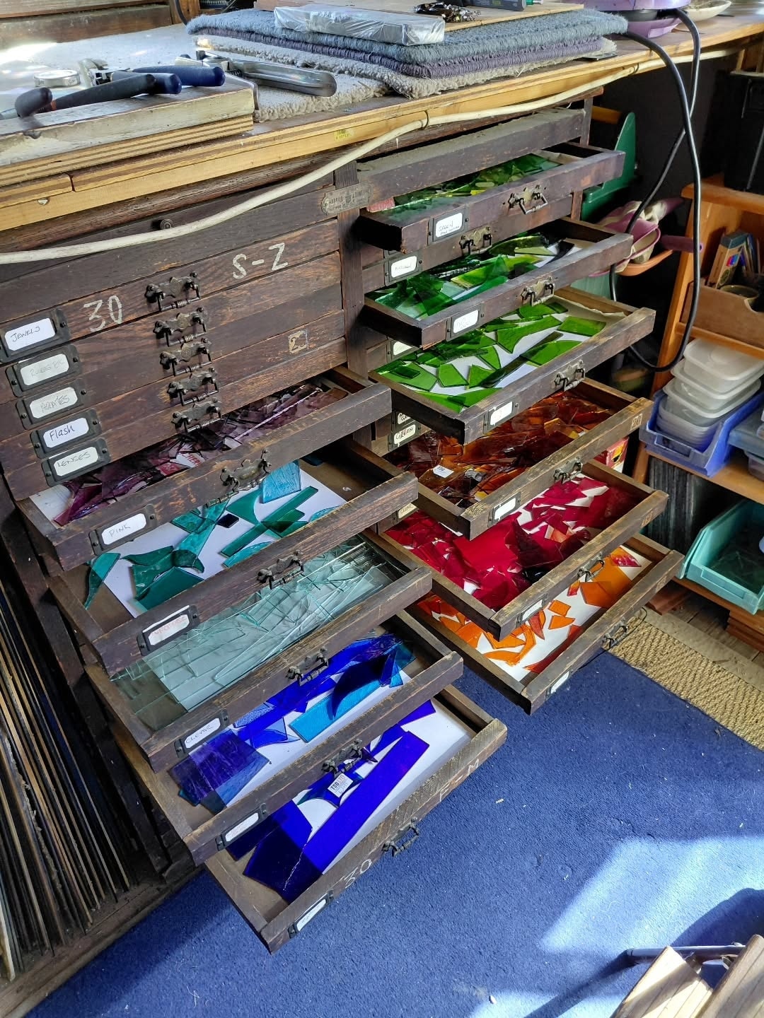 Shallow wooden drawers pulled open to show sheets of coloured glass sorted by colour