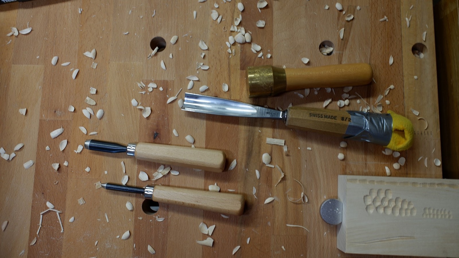 Gouges and pale wood shavings on a beech workbench beside a carved test block