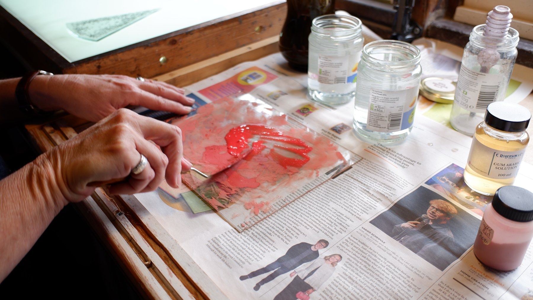 Hands spreading red glass paint across a sheet of glass on the bench