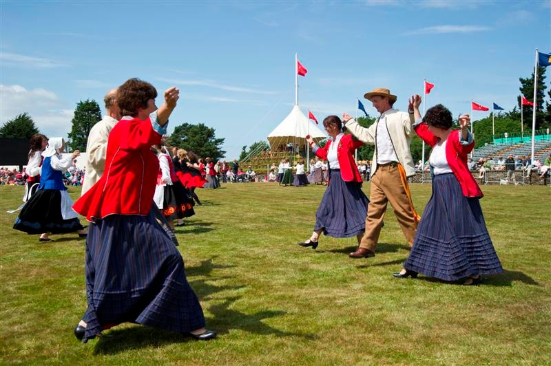 Performers in traditional red and blue costumes with flags dancing on grass at an outdoor festival, with a cream marquee and