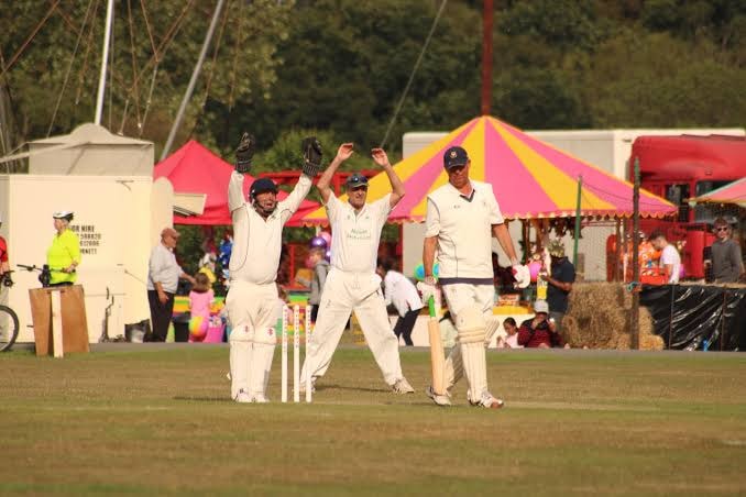 Cricket players in white uniforms celebrating on grass field, with colorful pink and yellow striped tent, spectators and boat