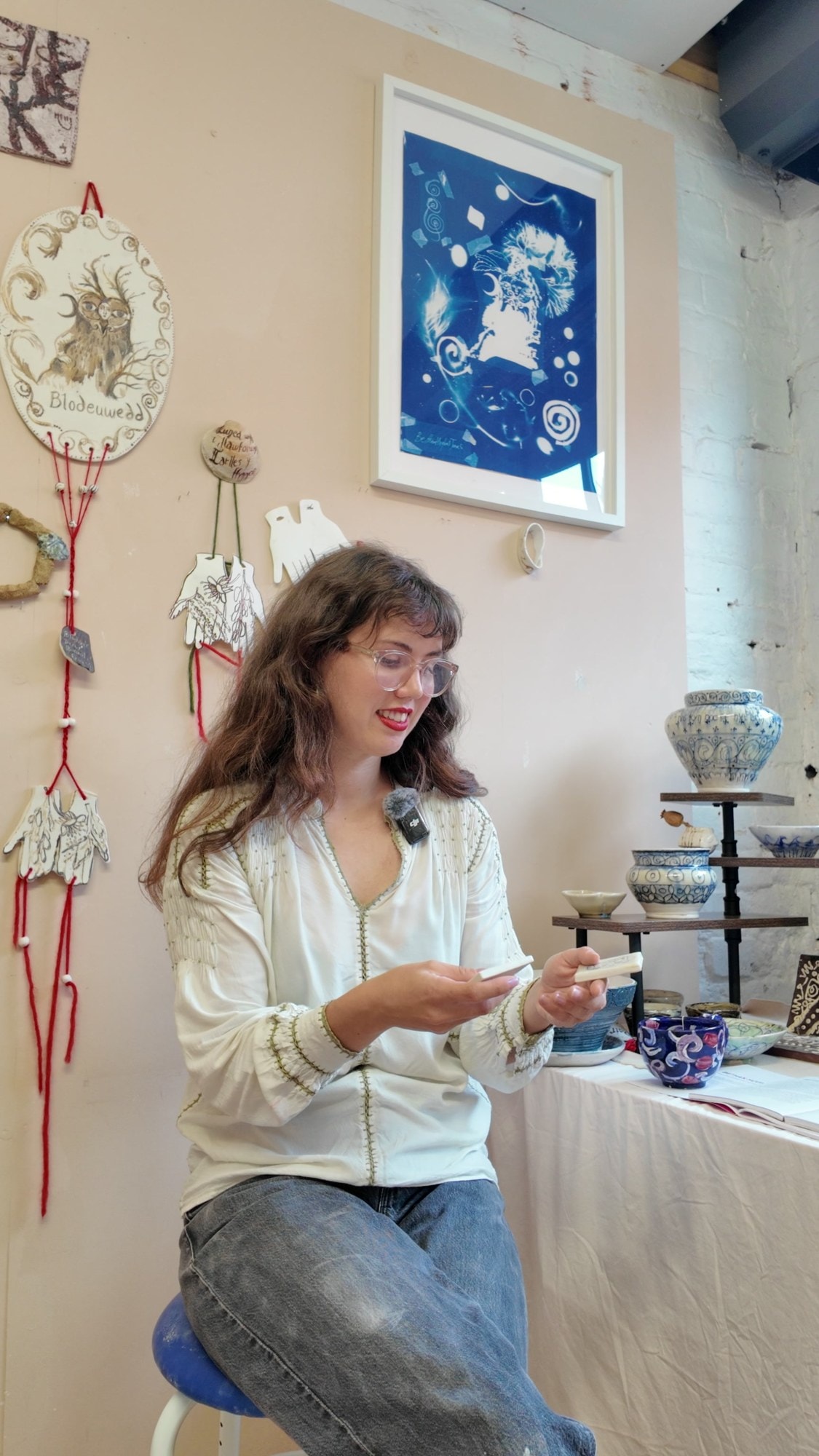 Bethan Hughes Jones smiling as she looks down at her tiles, sitting in her studio with a cyanotype print and her hanging ceramics behind
