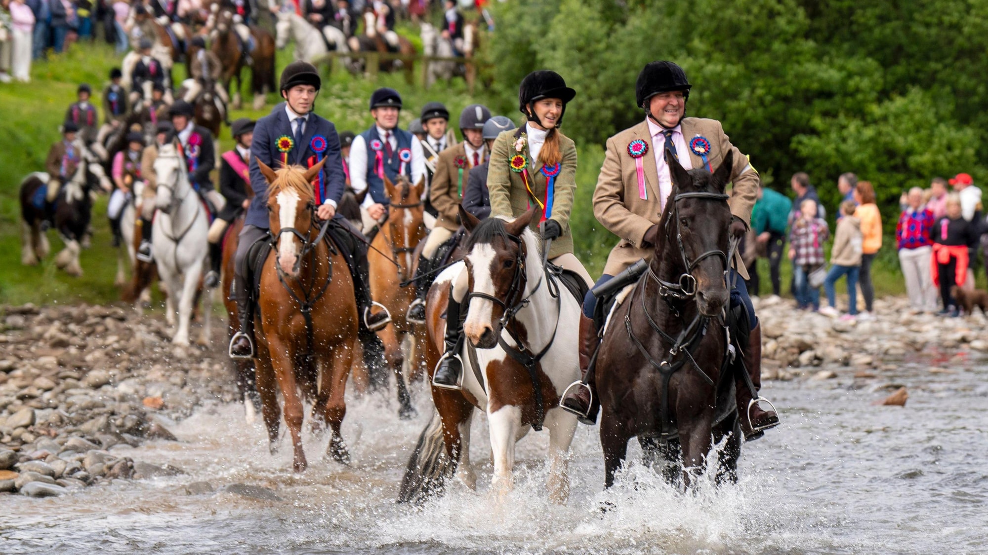 Riders in formal jackets and black helmets galloping through shallow water on horses of varying colours, wearing rosette ribb