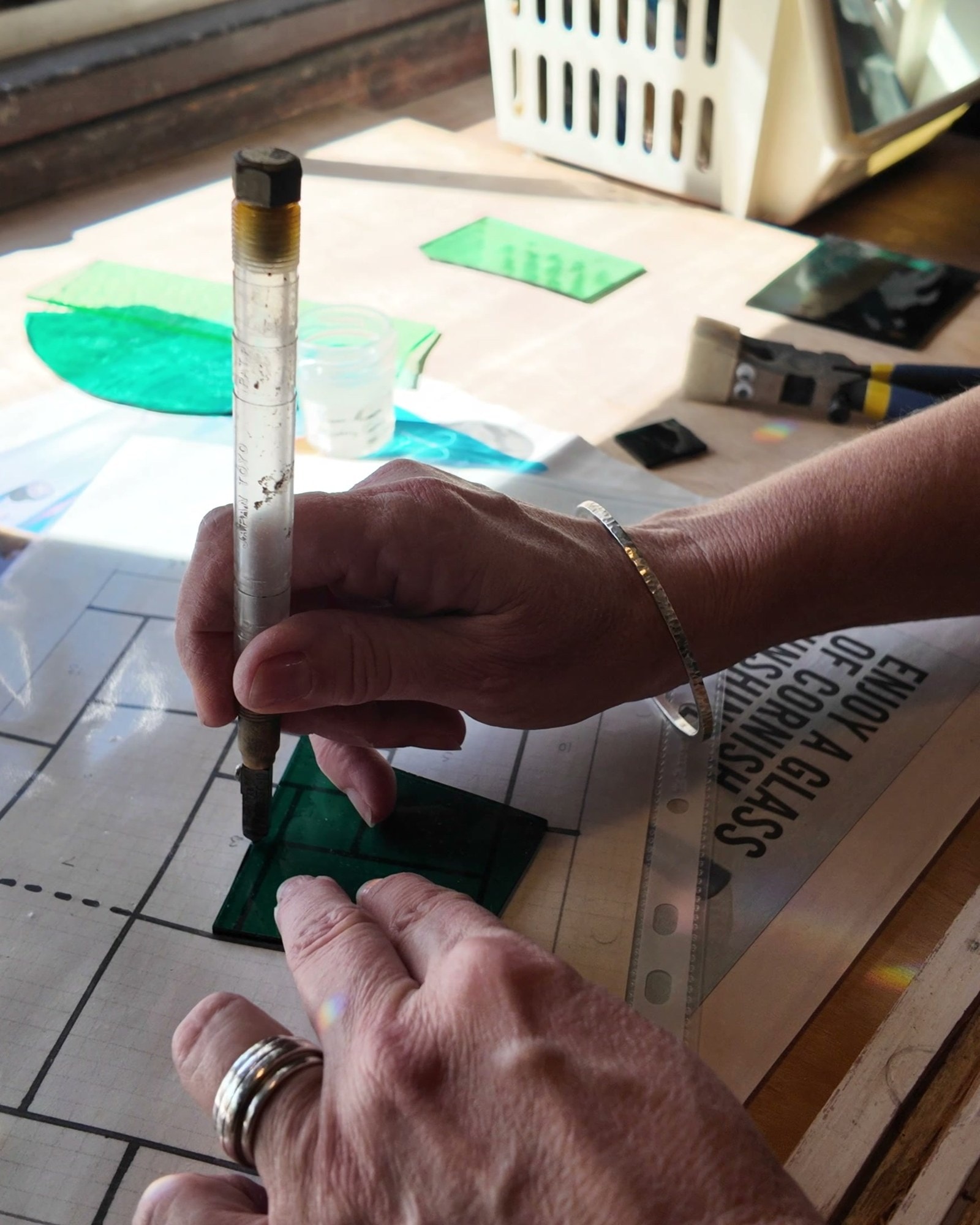 Mary Corkery's hands scoring a piece of green glass with a glass cutter over a drawing