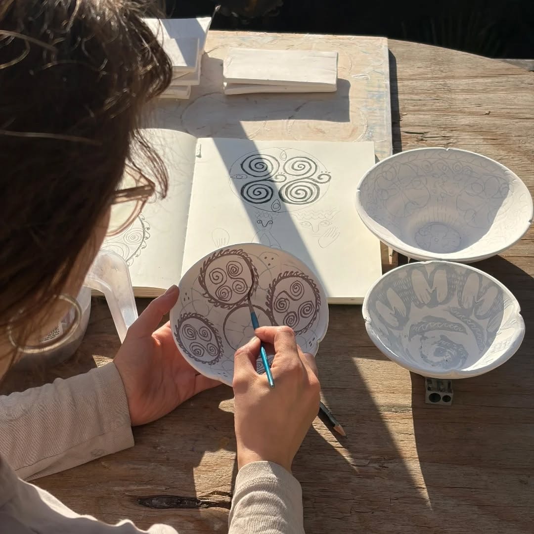 Hands decorating a porcelain disc at an outdoor bench beside a sketchbook and two bowls