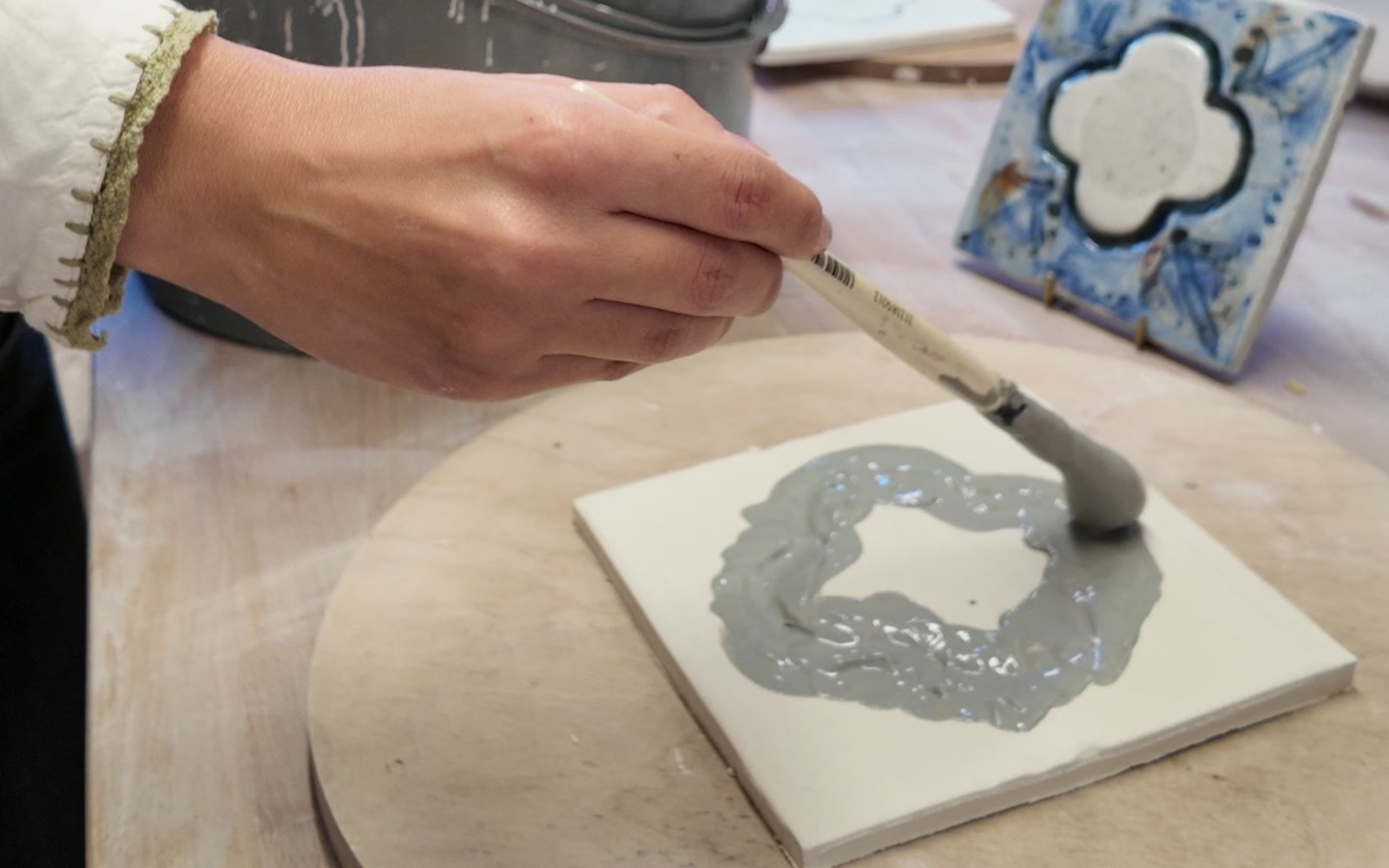 Bethan brushing grey slip into the stamped quatrefoil on a porcelain tile, a finished blue tile behind