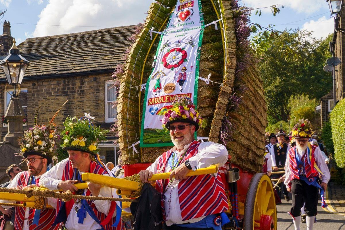 Men in traditional red and white striped waistcoats and flower crowns carrying yellow poles during a village procession, ston