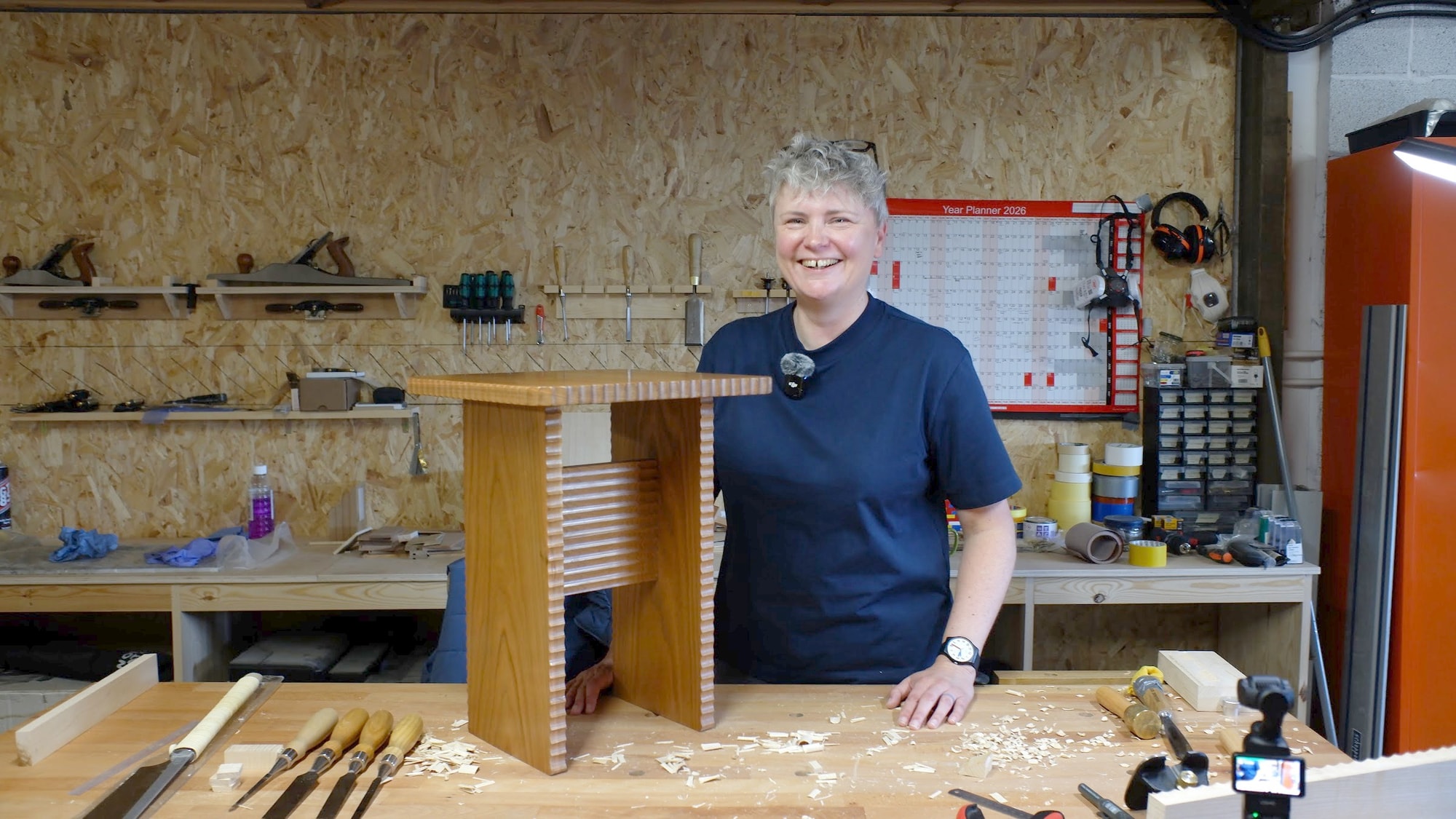 Claire Broad laughing behind a solid cherry side table with carved scalloped edges, in her workshop